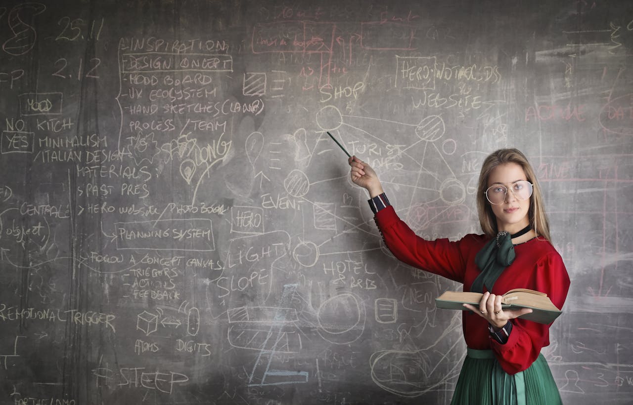 digital Serious female teacher wearing old fashioned dress and eyeglasses standing with book while pointing at chalkboard with schemes and looking at camera